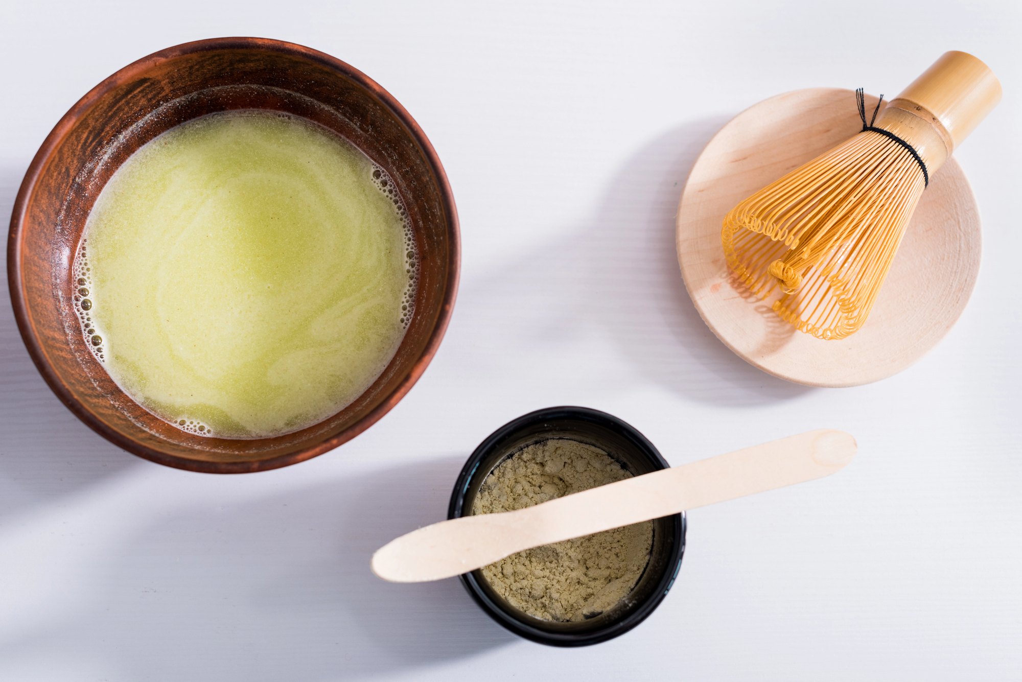 A bowl of prepared matcha tea, a bamboo whisk on a plate, and a container of matcha powder with a wooden spatula are arranged on a white surface, evoking the grace and precision of Japanese tea ceremony etiquette.