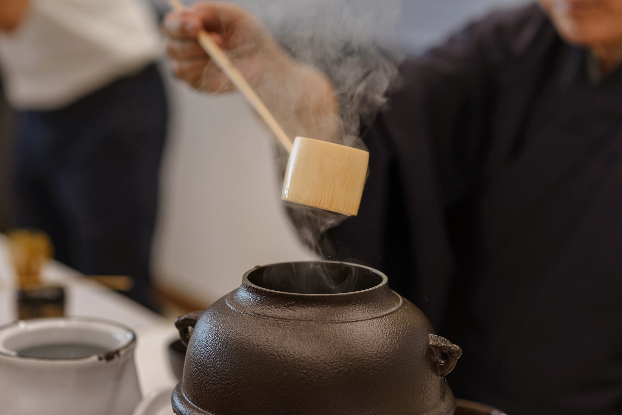 A person in traditional clothing pours hot water from a wooden ladle into a cast iron teapot, with steam rising—capturing the grace and precision of the Japanese tea ceremony.