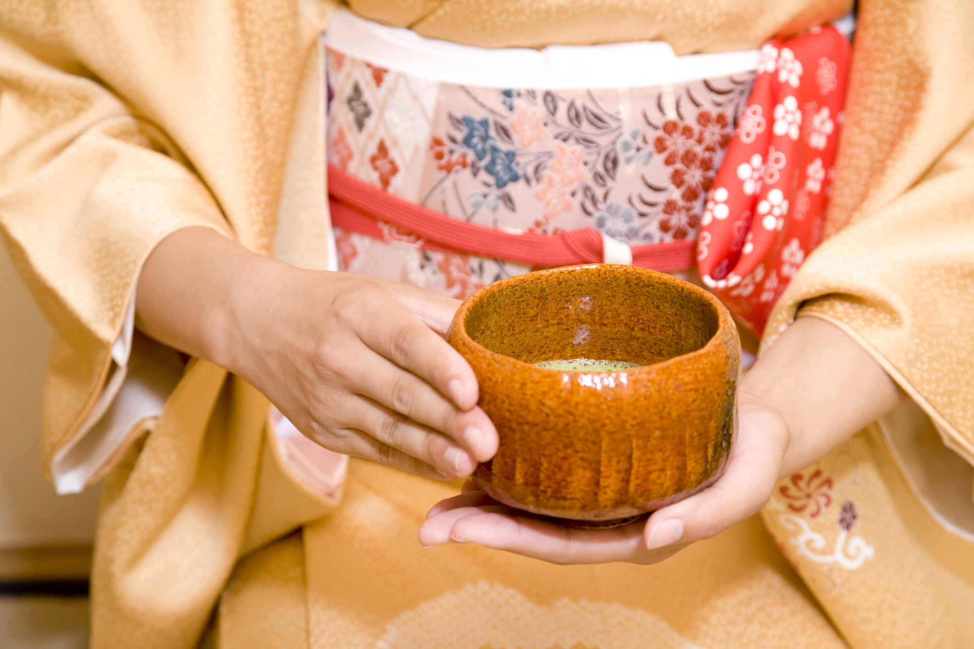 Person in a yellow patterned kimono holds a brown ceramic tea bowl with both hands, highlighting the etiquette central to the Japanese Tea Ceremony.