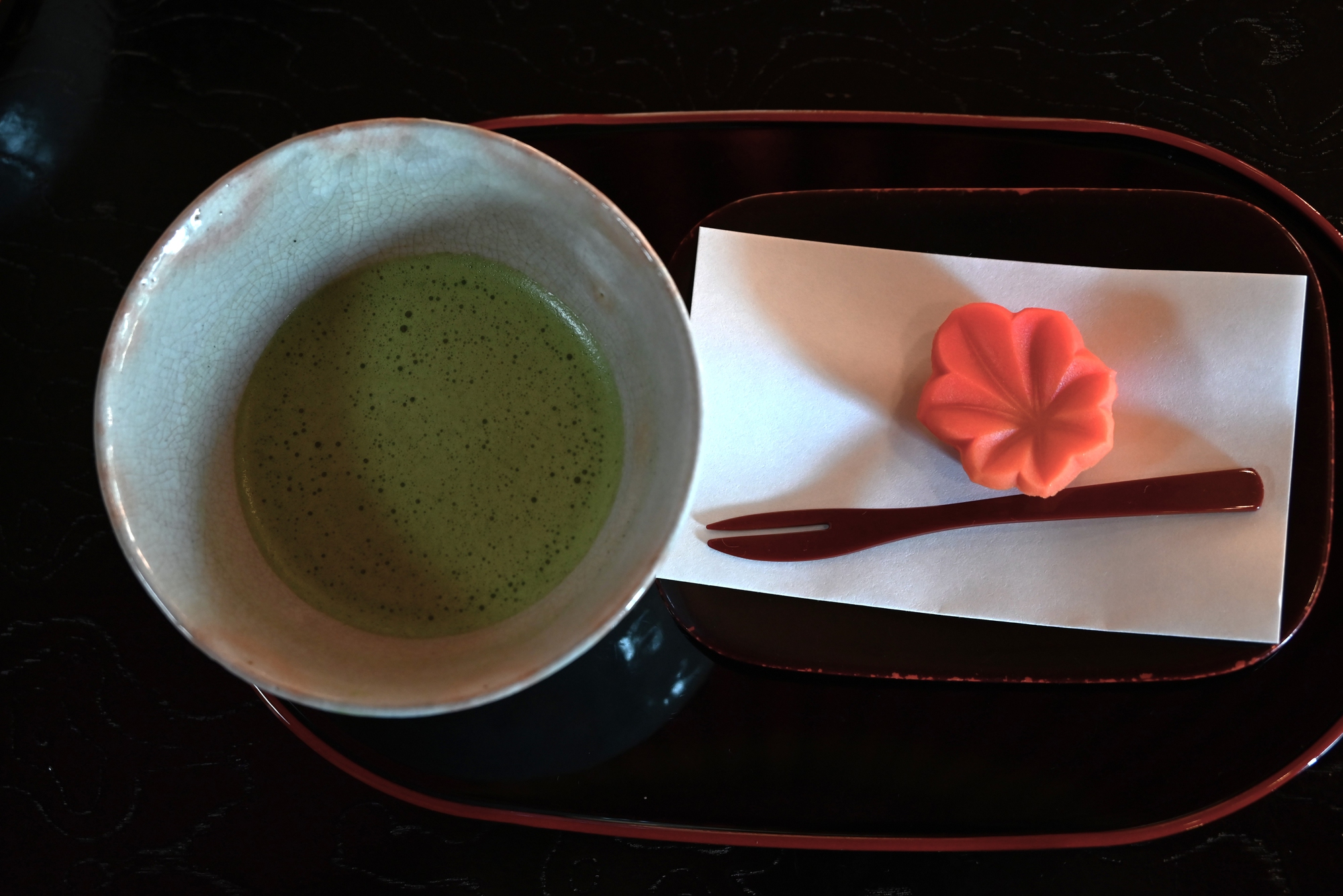 A cup of matcha green tea sits next to a pink flower-shaped wagashi sweet and a small fork on a tray with white paper, capturing the essence of Japanese Tea Ceremony and its refined tea ceremony etiquette.