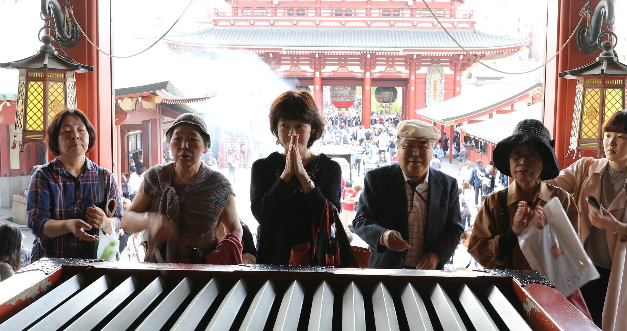 Five people stand at a Japanese temple altar behind an offering box; one woman prays with hands together, observing proper temple etiquette. Others stand nearby. A crowd and traditional architecture are visible in the background.