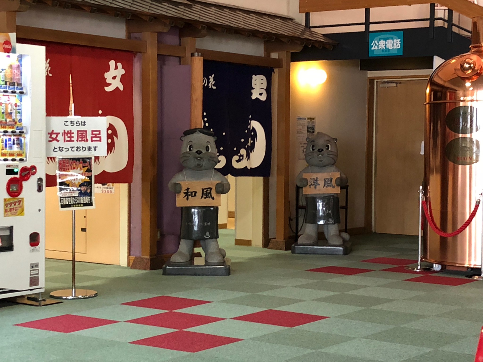 Two tanuki statues hold signs in front of curtained entrances labeled for men and women in a Japanese bathhouse, offering a glimpse of how to visit an onsen in Japan. A vending machine is on the left, with a copper object on the right.