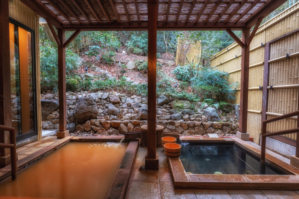Two outdoor Japanese hot spring baths with wooden buckets, surrounded by bamboo fencing, rocks, and lush greenery, under a wooden roof. The water is different colour in each bath due to mineral content.