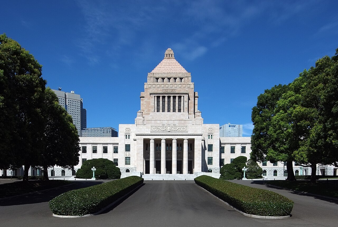 The National Diet Building in Tokyo, Japan, features a symmetrical façade and central pyramid-shaped tower, framed by green trees under a clear blue sky, offering a beautiful sight during May in Japan.