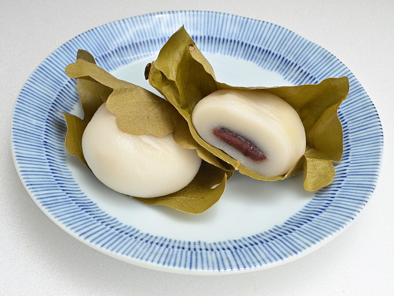 Two pieces of Japanese kasiwa mochi, a seasonal treat enjoyed in May in Japan, are wrapped in oak leaves on a blue and white plate; one is whole and the other cut open to reveal sweet red bean filling inside.
