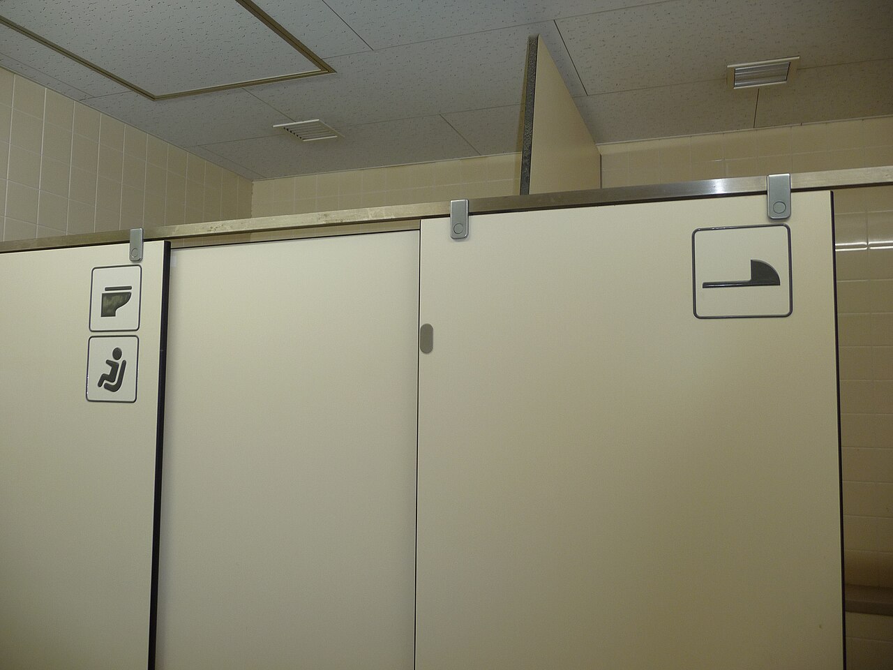 Photo taken inside a Japanese public toilet facility showing the top part of two bathroom stalls with pictograms; left stall has a squat toilet symbol, right stall has a Western toilet symbol. Beige tiled walls and ceiling visible above.