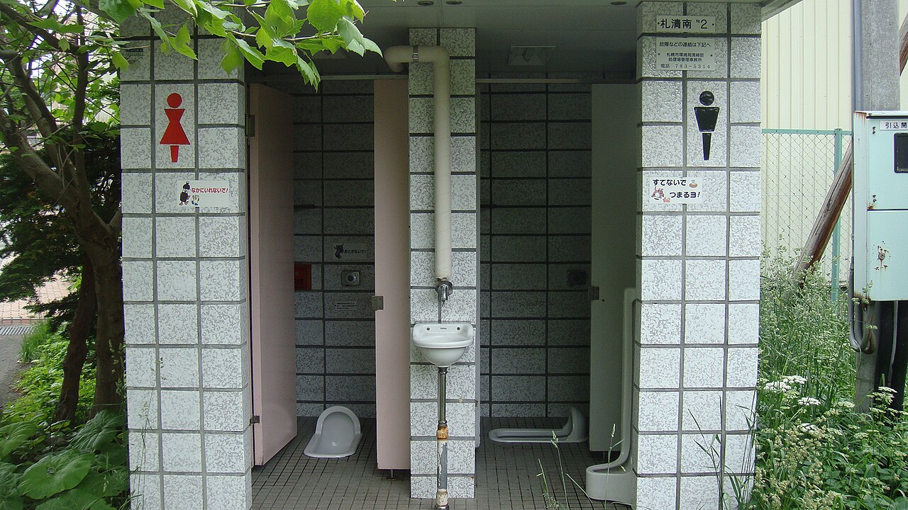 Outdoor public restroom with two stalls—one for women and one for men—tiled walls, a shared sink, Japanese signage. Two traditional Japanese style squat toilets are visible through the open doors.
