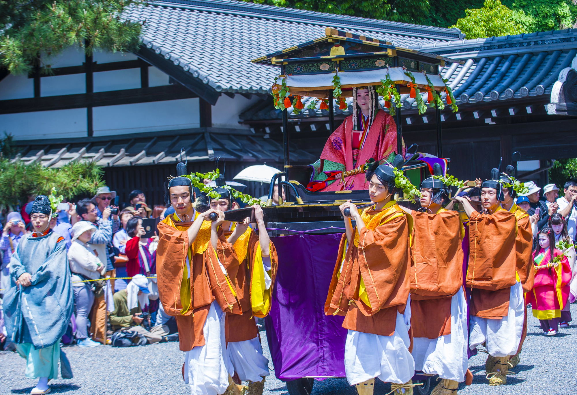 During an outdoor festival or parade, a group of people in traditional Japanese clothing carry a decorated palanquin with a person inside, celebrating customs often seen in May in Japan.