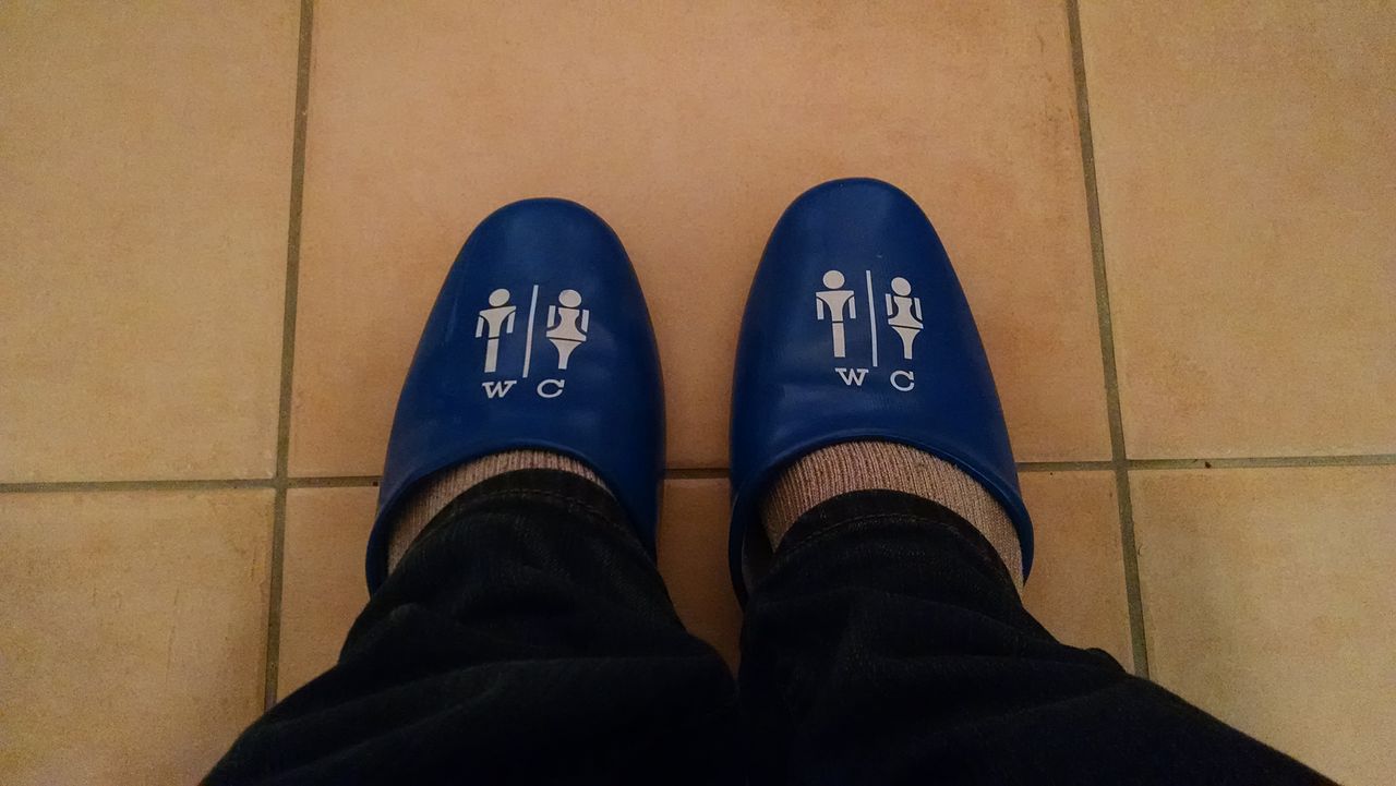 A person wearing blue Japanese toilet slippers labeled "WC" with male and female restroom symbols stands on a tiled floor, ready to use a Japanese toilet.