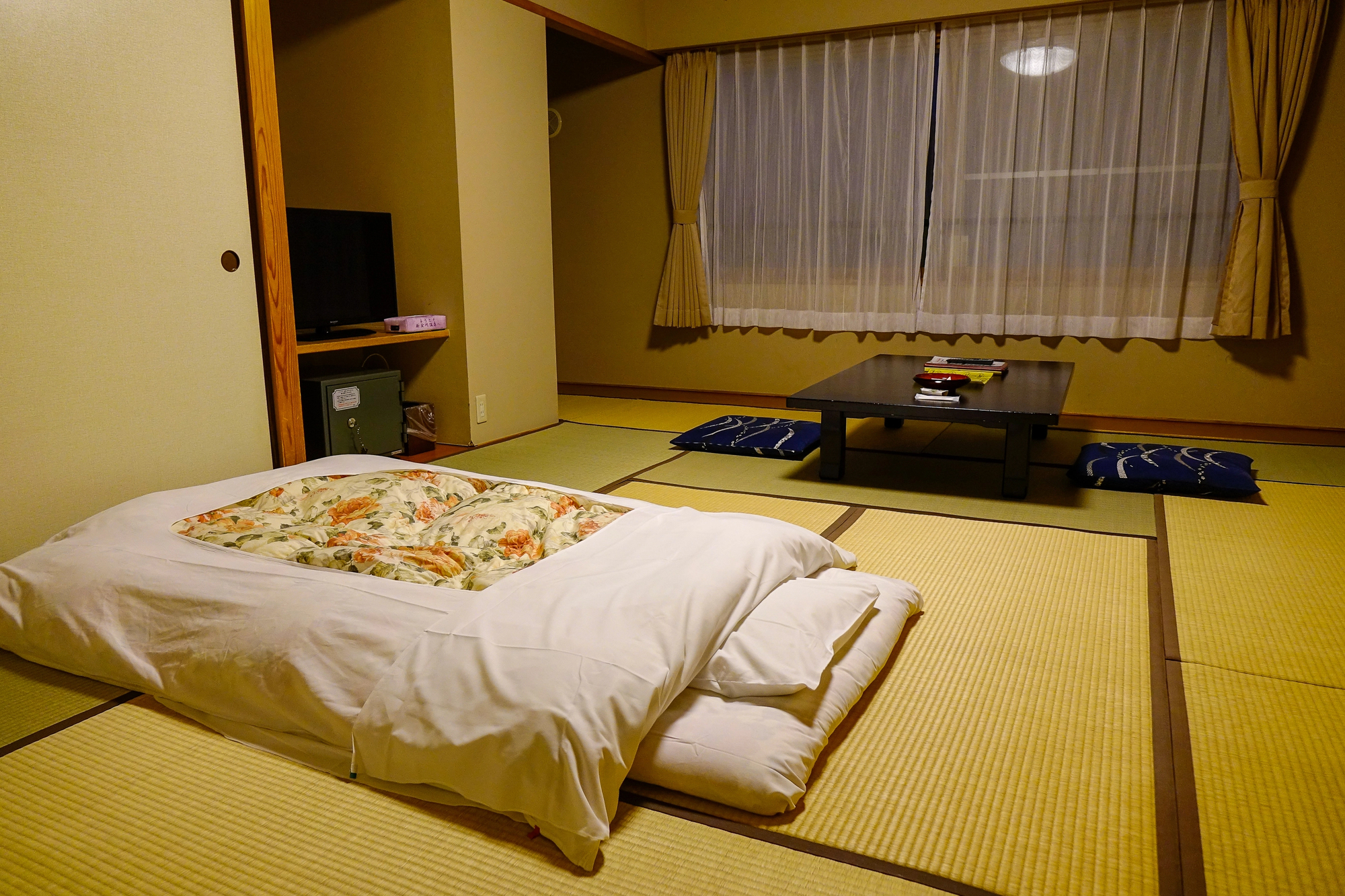 A traditional Japanese Ryokan room with tatami flooring, a futon bed, a low table with floor cushions, a window with curtains, and a television in the corner.
