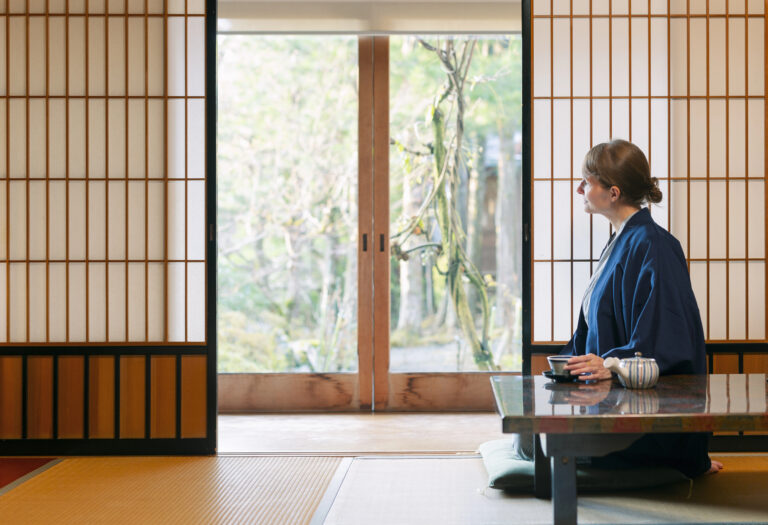 A person in traditional Japanese clothing sits at a low table with tea, enjoying the serene garden view through sliding shoji doors—capturing the essence of an authentic Japanese Ryokan experience.
