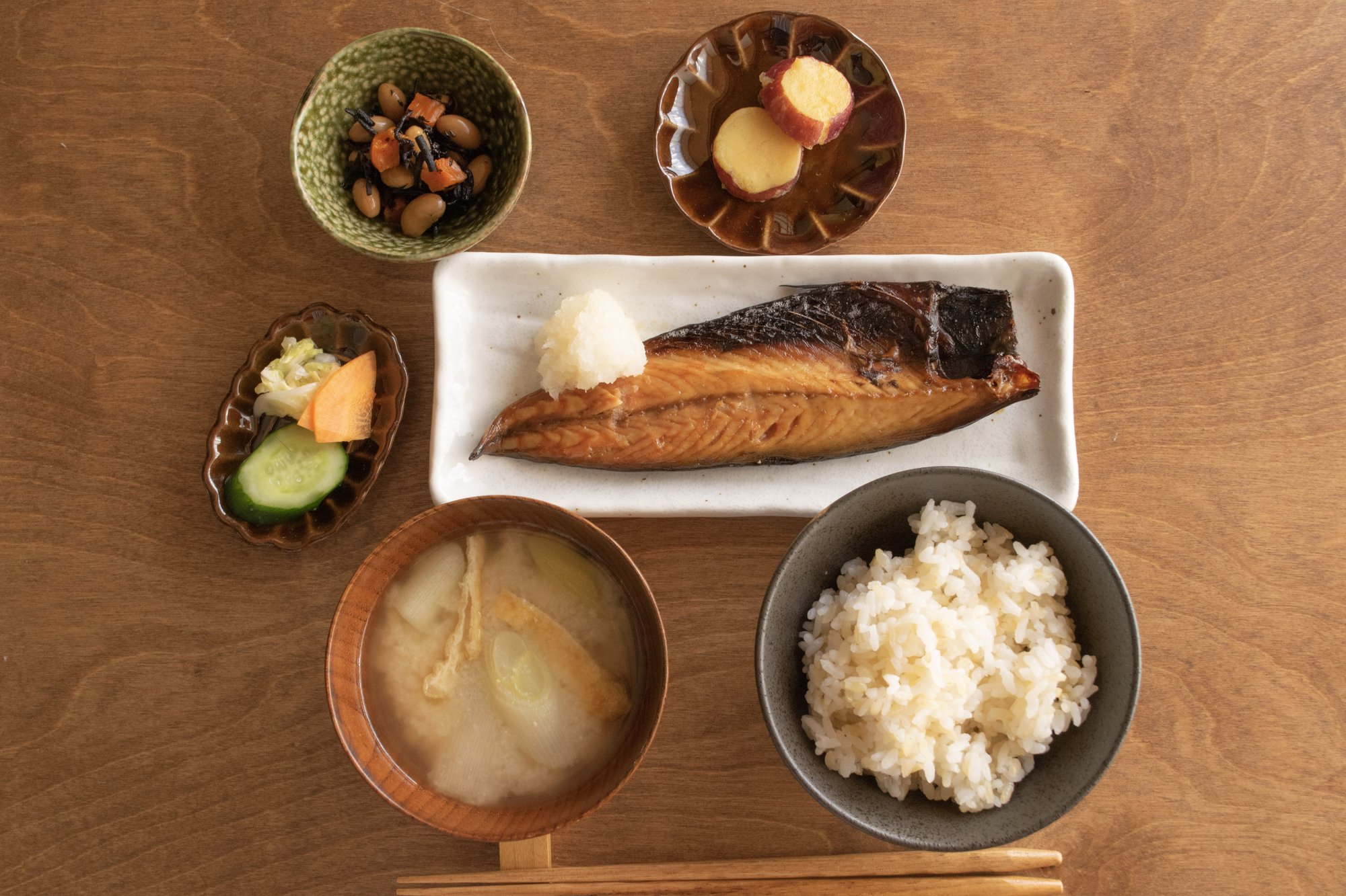 A Japanese traditional breakfast meal featuring grilled fish, white rice, miso soup, pickled and simmered vegetables, and a small salad is beautifully arranged on a wooden table with chopsticks, offering an authentic ryokan experience.