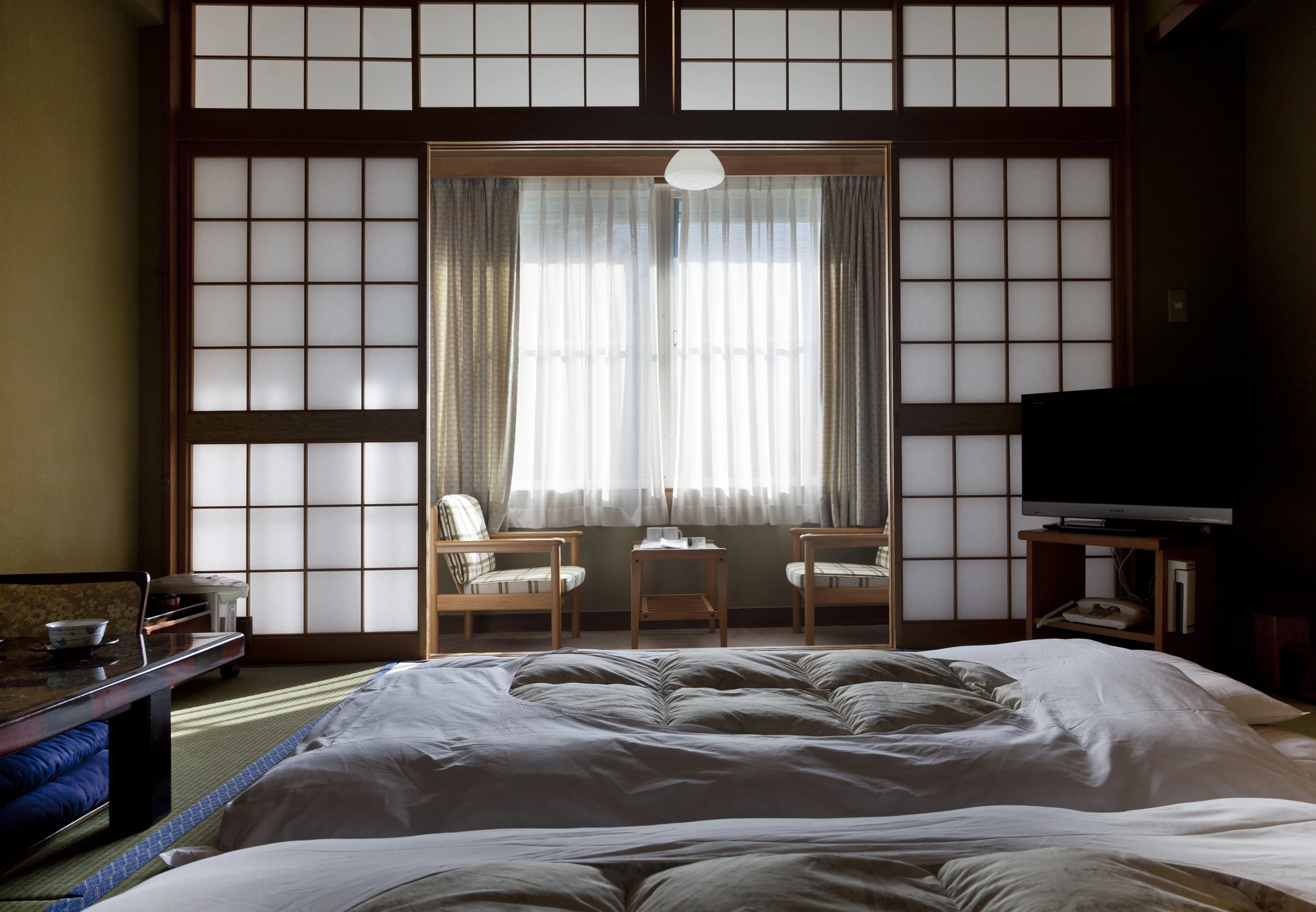 A traditional Japanese ryokan room with shoji screens, a low table, two chairs by the window, a television, and a futon bed on tatami flooring offers an authentic Japanese inn experience.