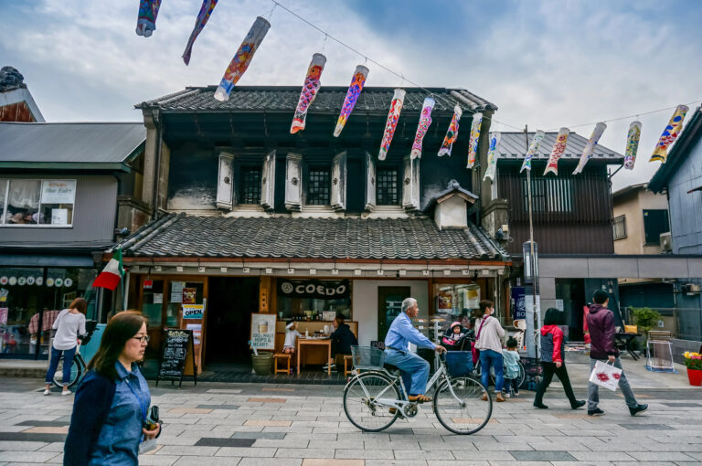 People walk and cycle along a street lined with traditional Japanese buildings, with colorful carp streamers hanging above for Children's Day on 5 May, capturing the festive spirit of May in Japan.