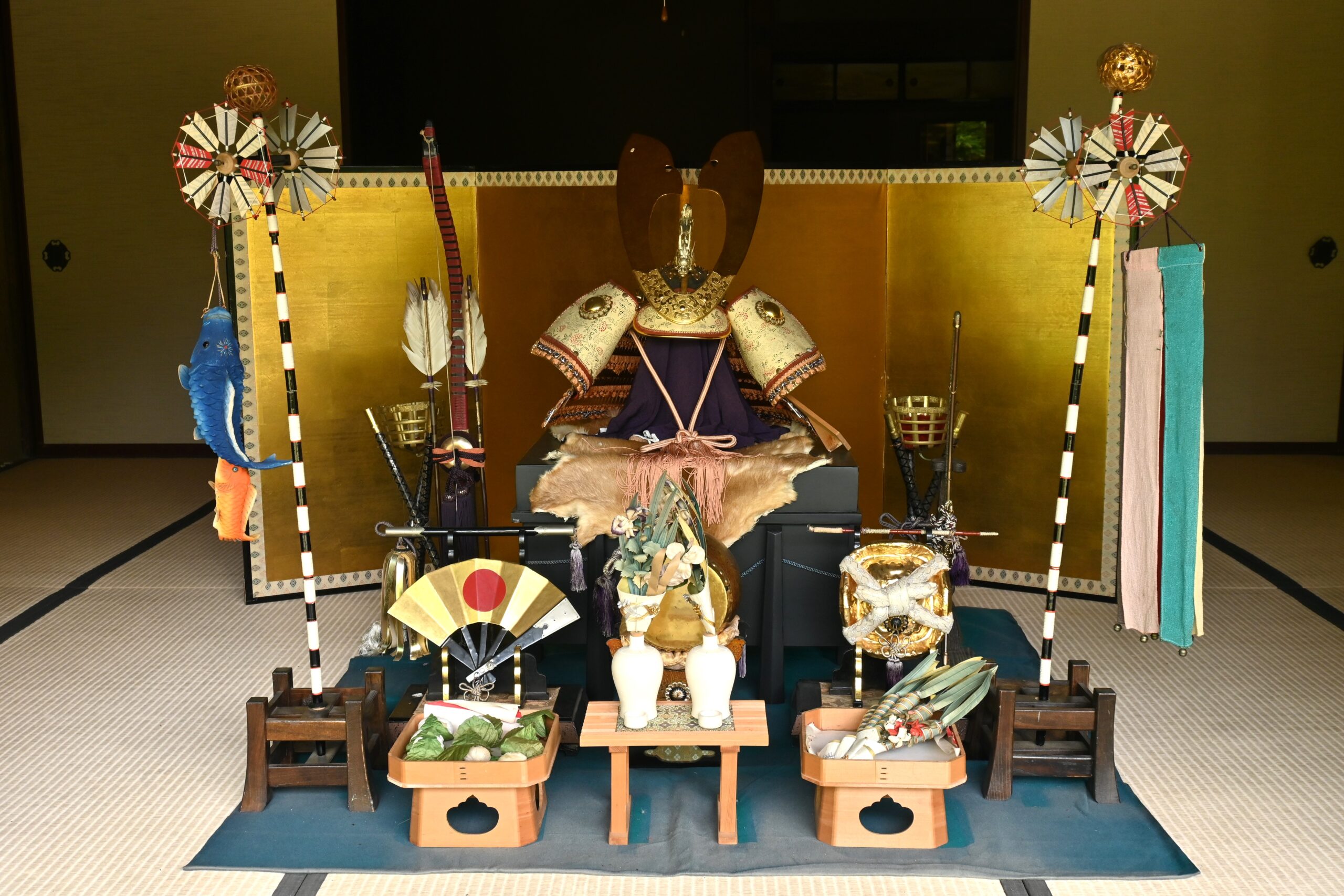 A traditional Japanese samurai armor display with helmets, weapons, golden screen, fans, and offerings arranged on a mat in an indoor setting—an impressive sight often seen during May in Japan.