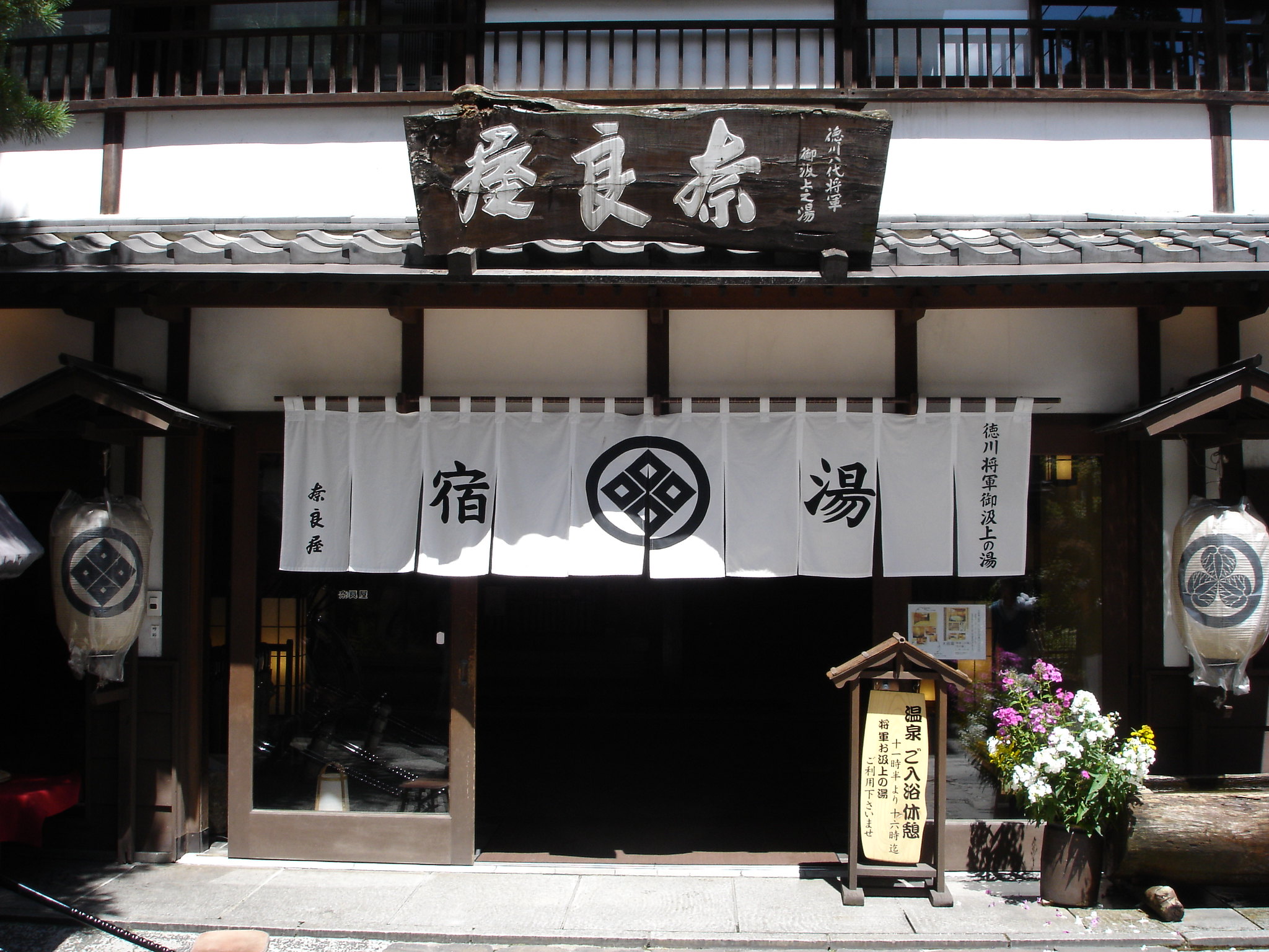 Traditional Japanese ryokan entrance with a white noren curtain featuring black symbols, wooden sign above, and flower arrangement beside a wooden information board.