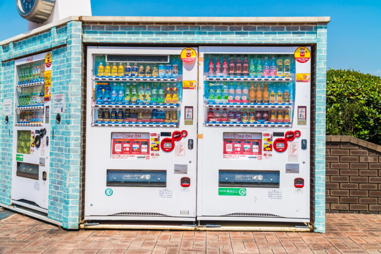 Two outdoor Japanese vending machines filled with assorted drinks stand inside a blue-tiled wall on a sunny day.