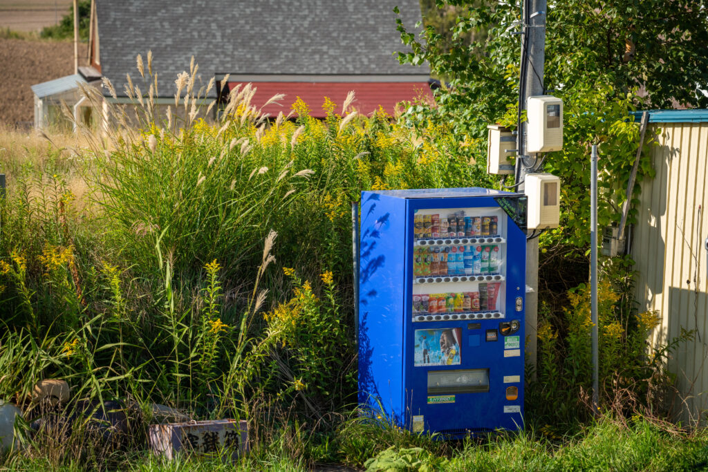 Japanese Vending Machine in countryside A blue Japanese vending machine stands outdoors in rural Japan surrounded by tall grass and weeds, with buildings and a field in the background.