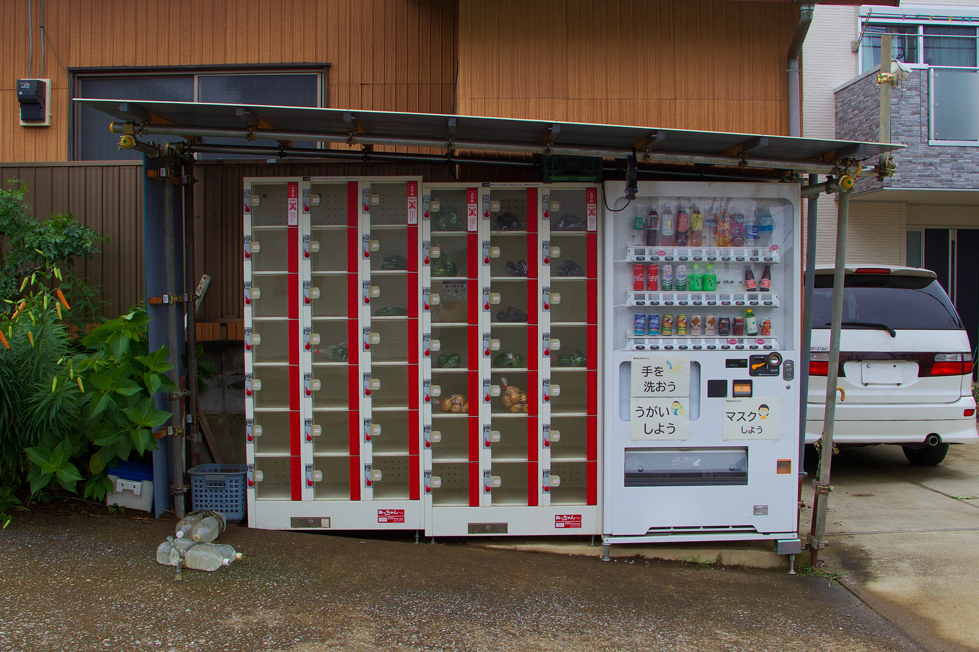 Japanese Vending Machine vegetables A Japanse vending machine selling drinks, with another vending machine next to it that consists of rows of small lockers with see-through doors, through which bags of vegetables are visible. They are situated under a metal awning next to a house and parked white vehicle.