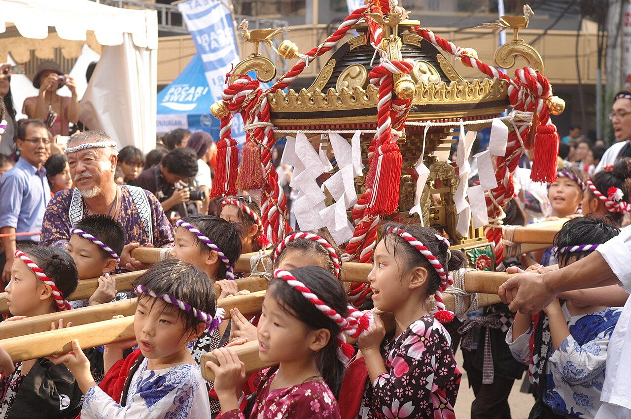 mikoshi A group of children in traditional Japanese summer festival clothing carry a decorated portable shrine (mikoshi) during a festival parade, with adults supervising nearby.