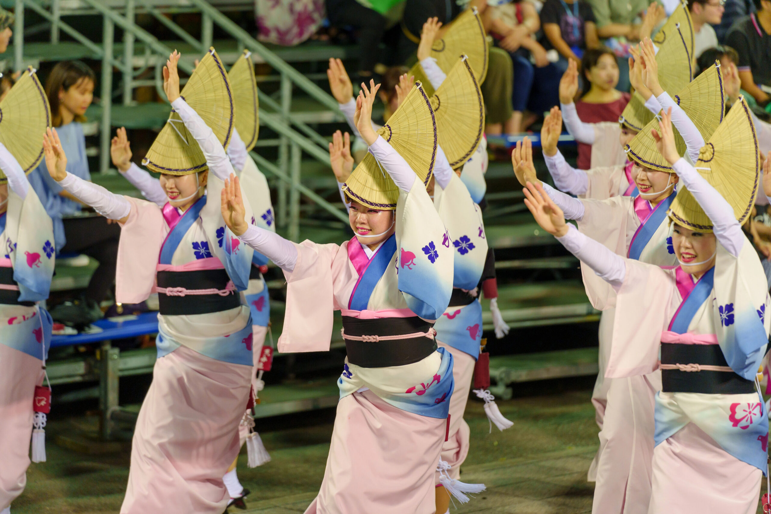 august in japan awa odori A group of women in traditional Japanese festival attire with amigasa straw hats perform a synchronized awa odori dance for an audience on tiered seating.
