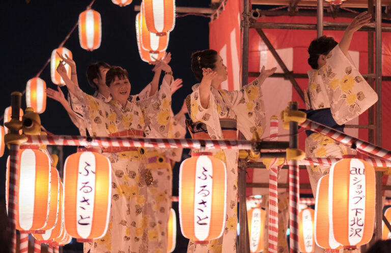 Women in yukata dance on a raised platform surrounded by illuminated paper lanterns at a nighttime obon dance festival held during August in Japan.