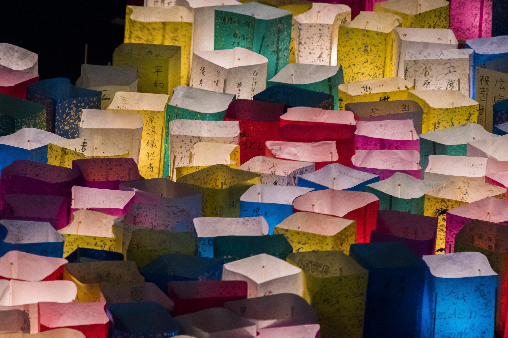 august in japan obon lanterns floating A collection of colorful paper lanterns with handwritten messages and characters, illuminated from within and arranged closely together, floating on a dark river at night.