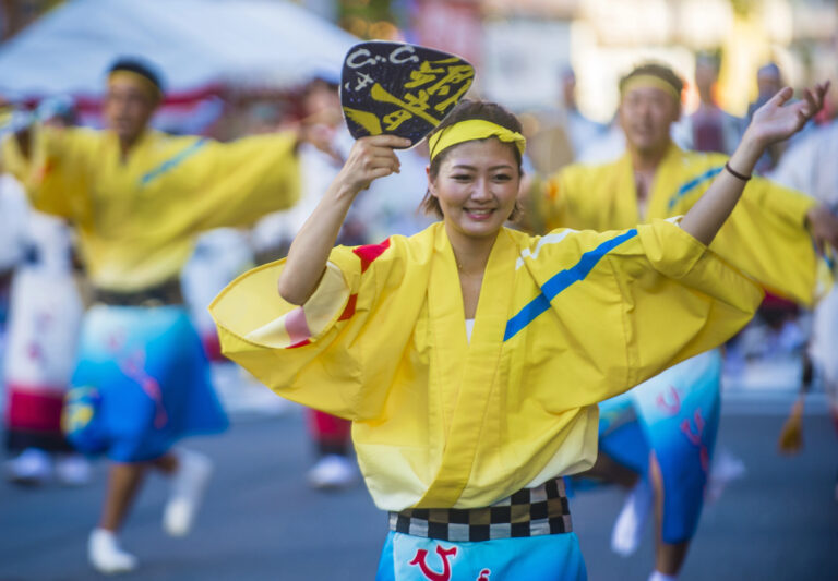 A woman in a yellow traditional Japanese festival outfit dances with a fan during a colourful summer street festival, with other performers in similar attire in the background.