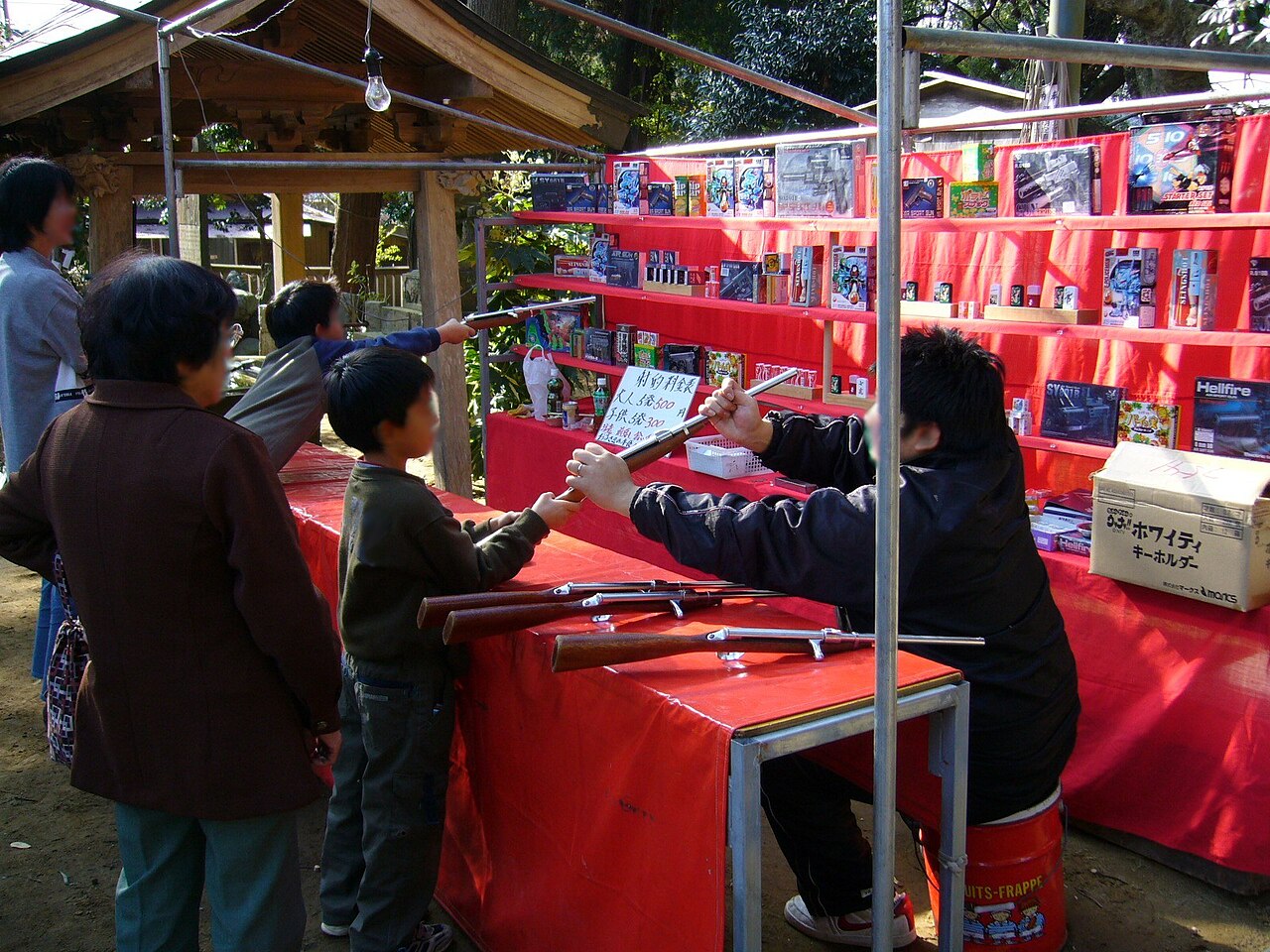japanese summer festival Shooting_gallery game,shateki,katori-city,japan Children and adults stand at a carnival-style shooting game booth with toy rifles and prizes displayed on red shelves.