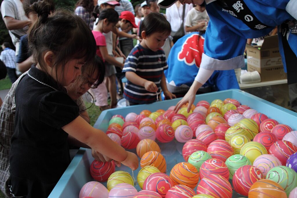 japanese summer festival balloon game Children select colorful floating balls from a water-filled tray at a festival booth, with adults and other kids standing nearby in the background.