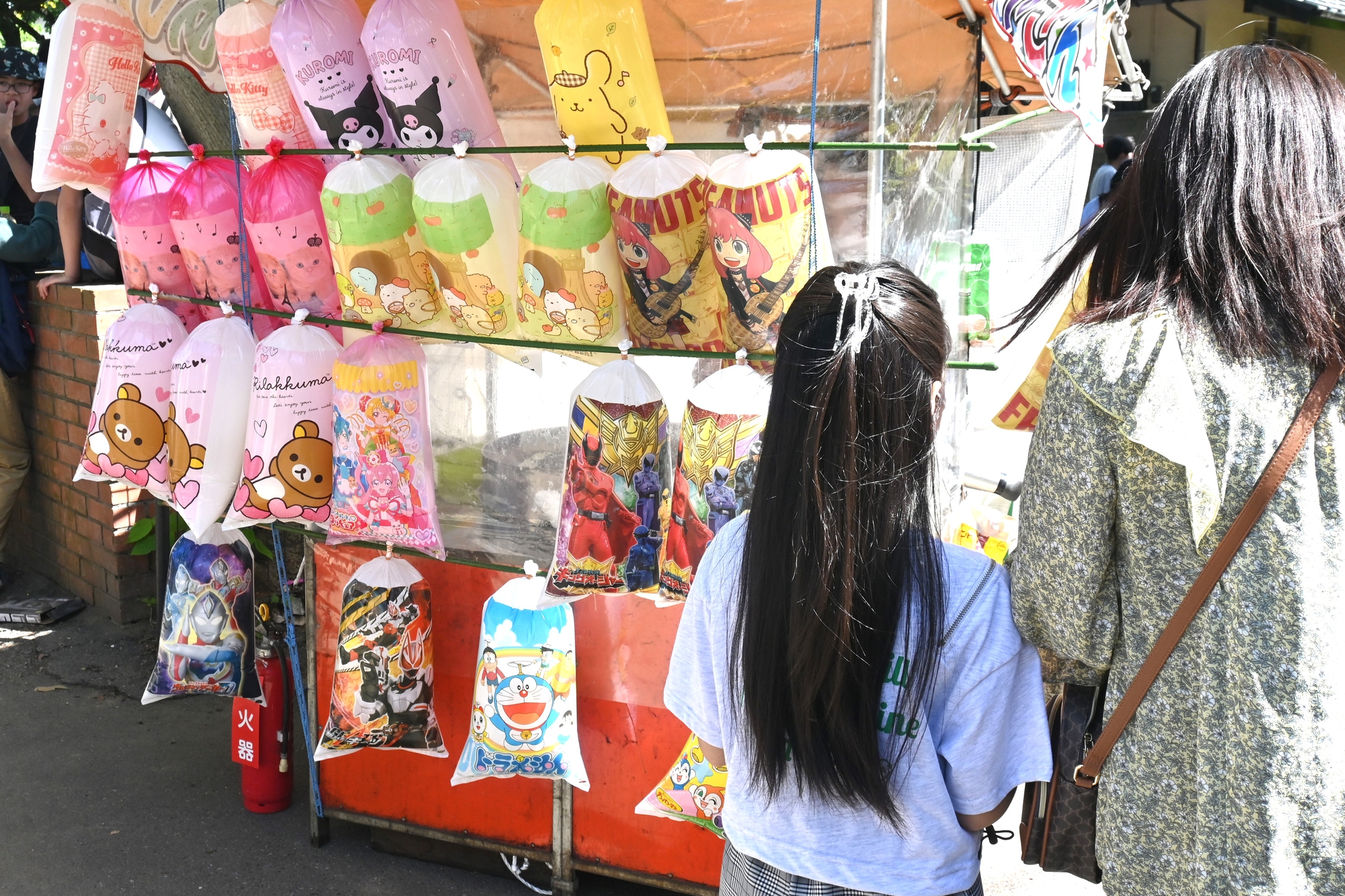 japanese summer festival cotton candy Two people stand in front of a street stall at a Japanese summer festival, admiring colorful bags of cotton candy featuring various cartoon characters.