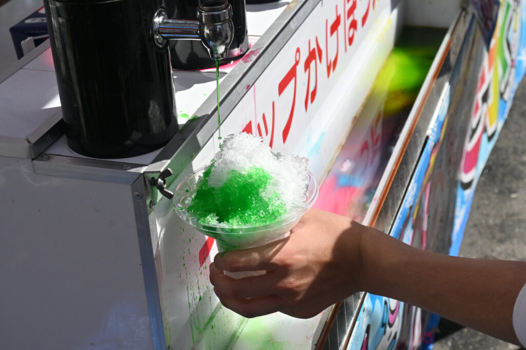 japanese summer festival shaved ice At a Japanese Summer Festival, a person pours green syrup over a cup of shaved ice from a dispenser at an outdoor stand.