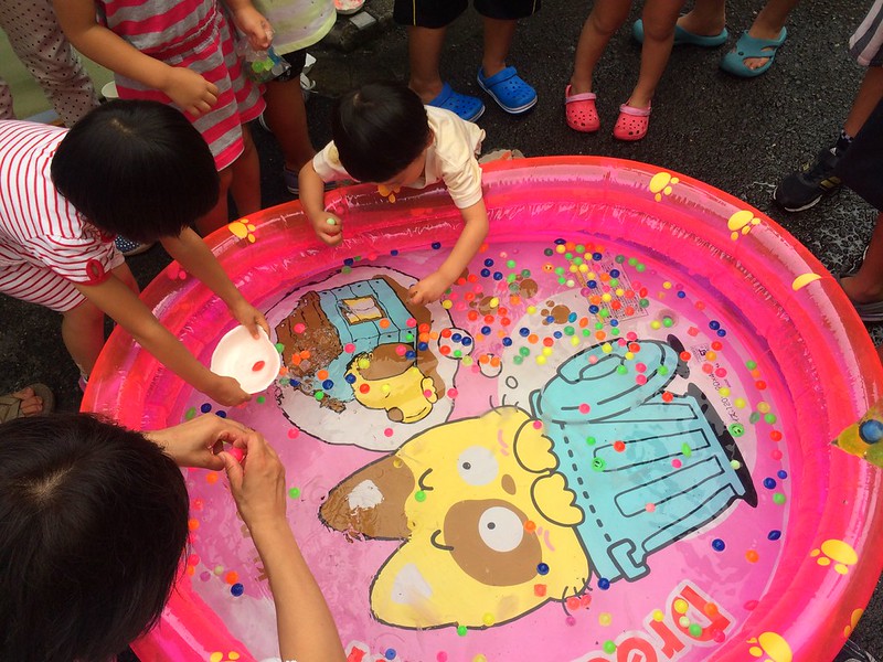 japanese summer festival super ball game Children gather around a pink inflatable pool, using small bowls and scoops to collect colorful floating beads.