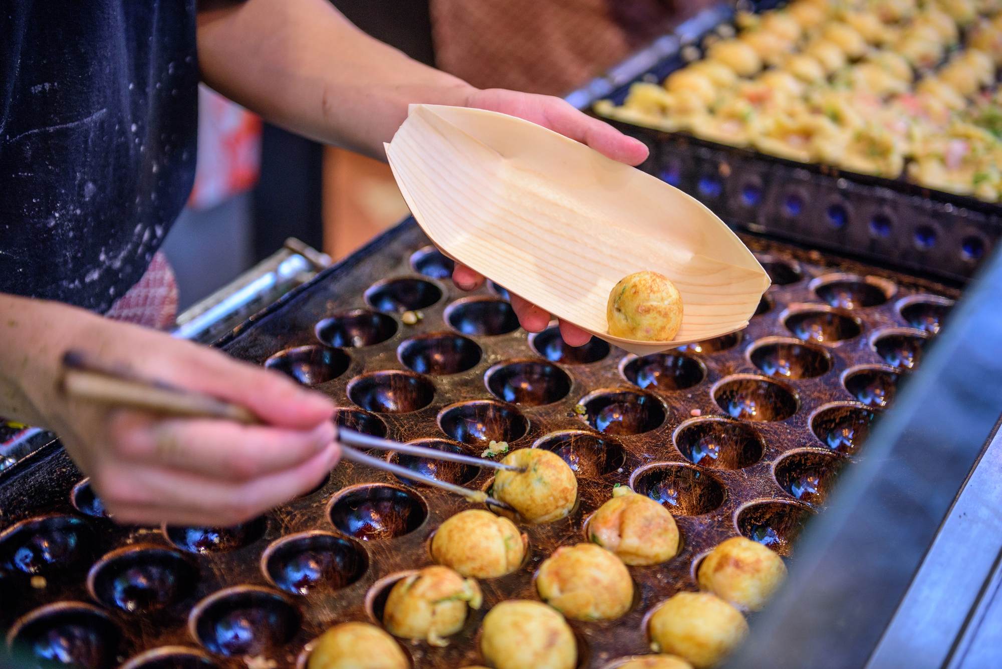 japanese summer festival takoyaki A person uses chopsticks to place freshly cooked takoyaki balls into a wooden serving tray at a takoyaki stand.