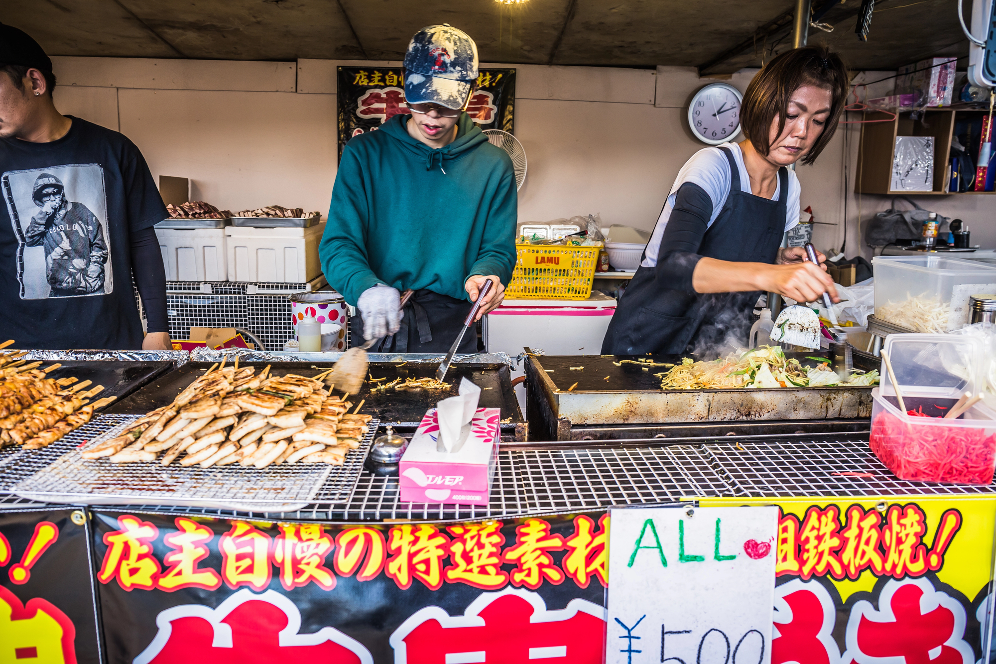 japanese summer festival yatai street food Two people cook and serve food at a Japanese street food stall at a summer festival, grilling yakitori skewers and yakisoba noodles on flat griddles with a price sign in front.