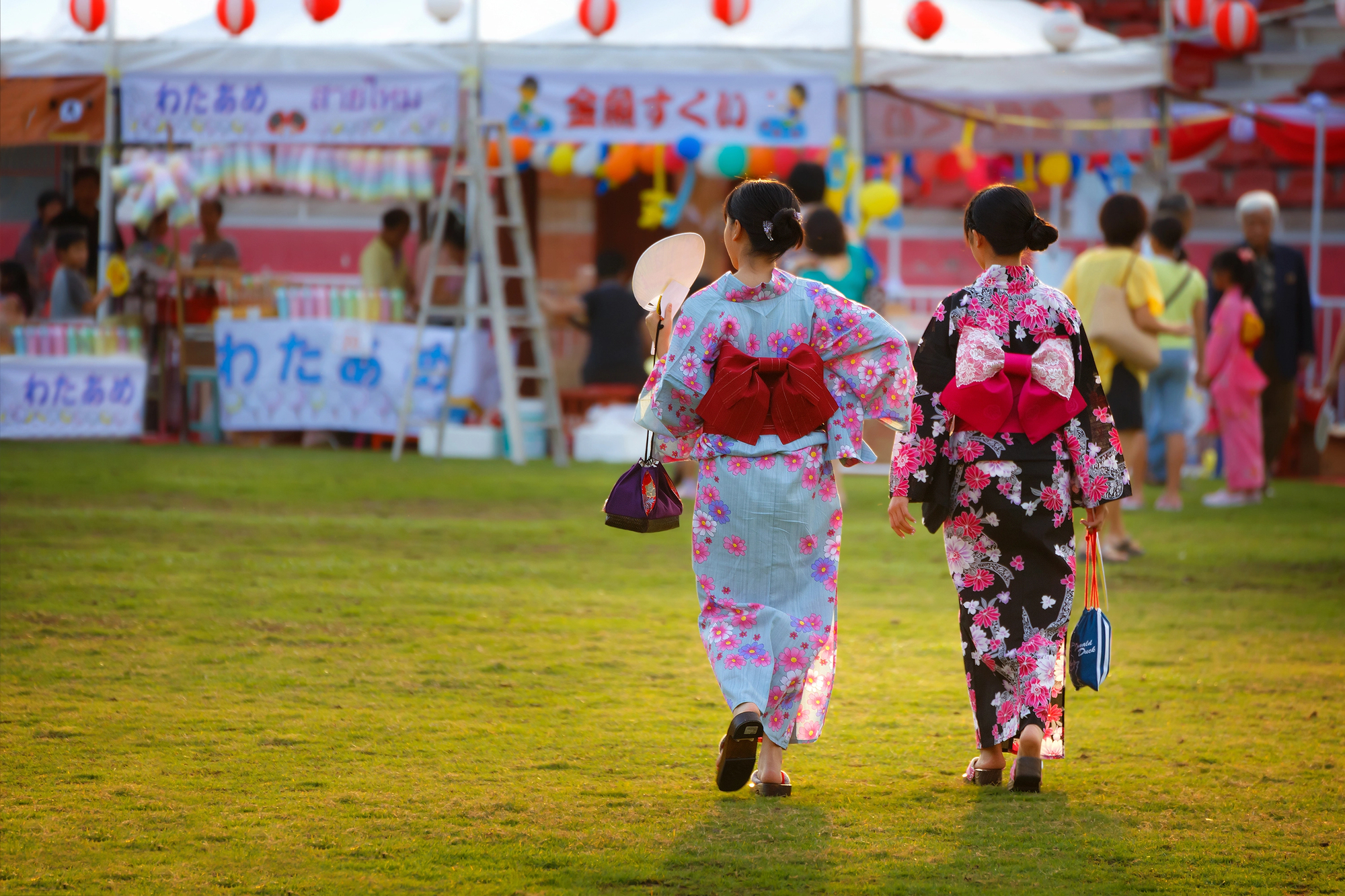 japanese summer festival yukata Two people wearing floral yukatas walk together at a Japanese summer festival, with food stalls and colorful decorations visible in the background.