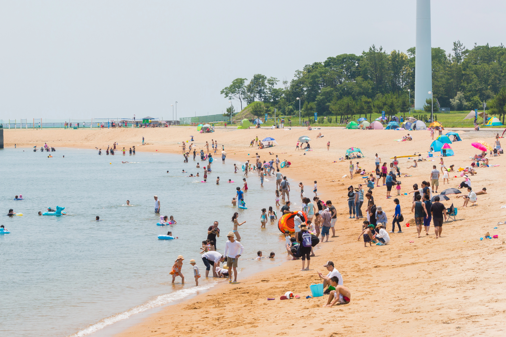 july in japan beach marine day Crowds of people enjoy swimming, playing, and relaxing on a sandy beach by the water during July in Japan, with umbrellas, tents, and trees in the background.