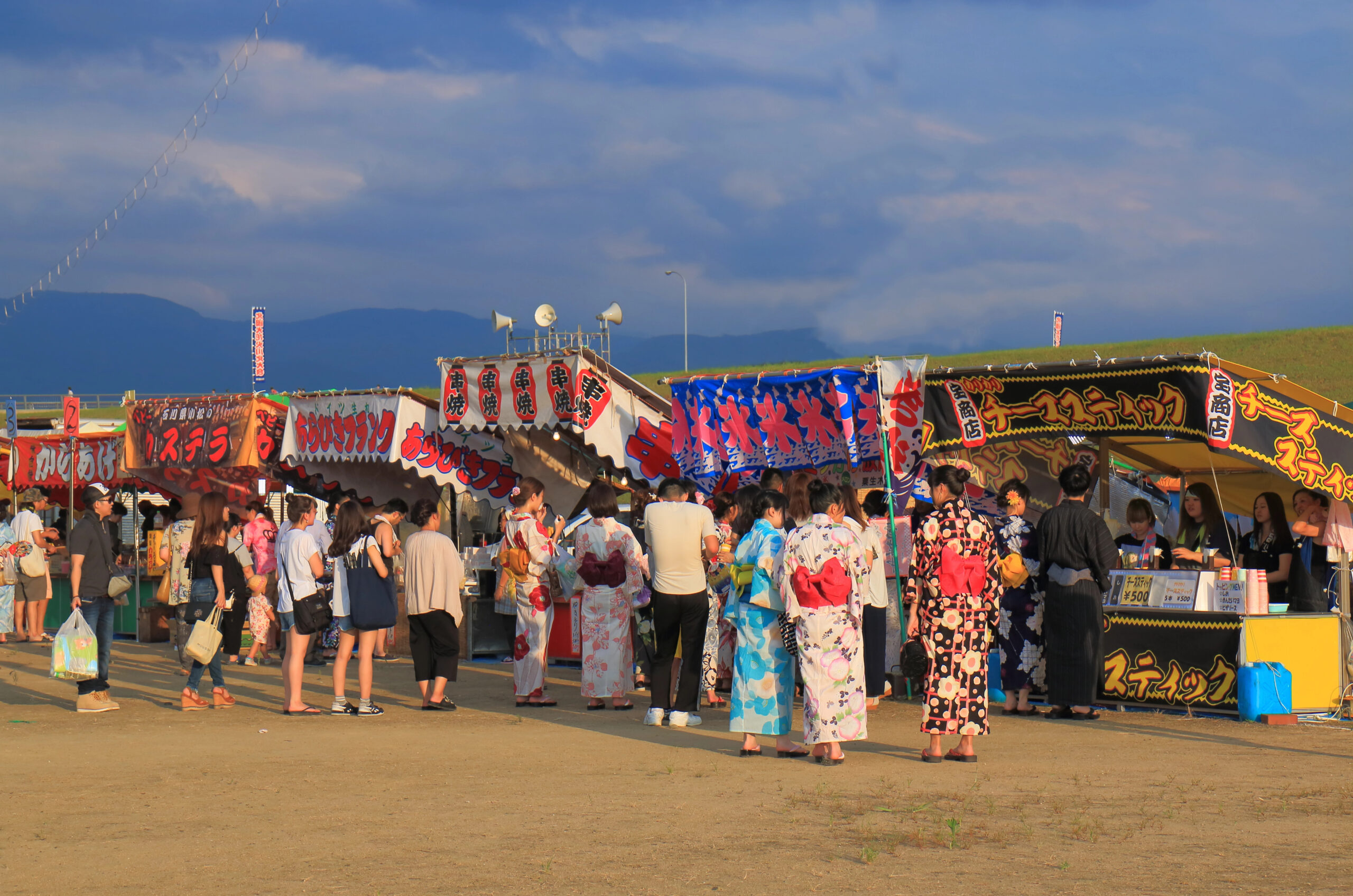 july in japan summer feestival yukata People in traditional yukata summer festival clothing stand in line at outdoor food stalls under colorful banners during a lively Japanese summer festival. The sky is partly cloudy and the ground is unpaved.