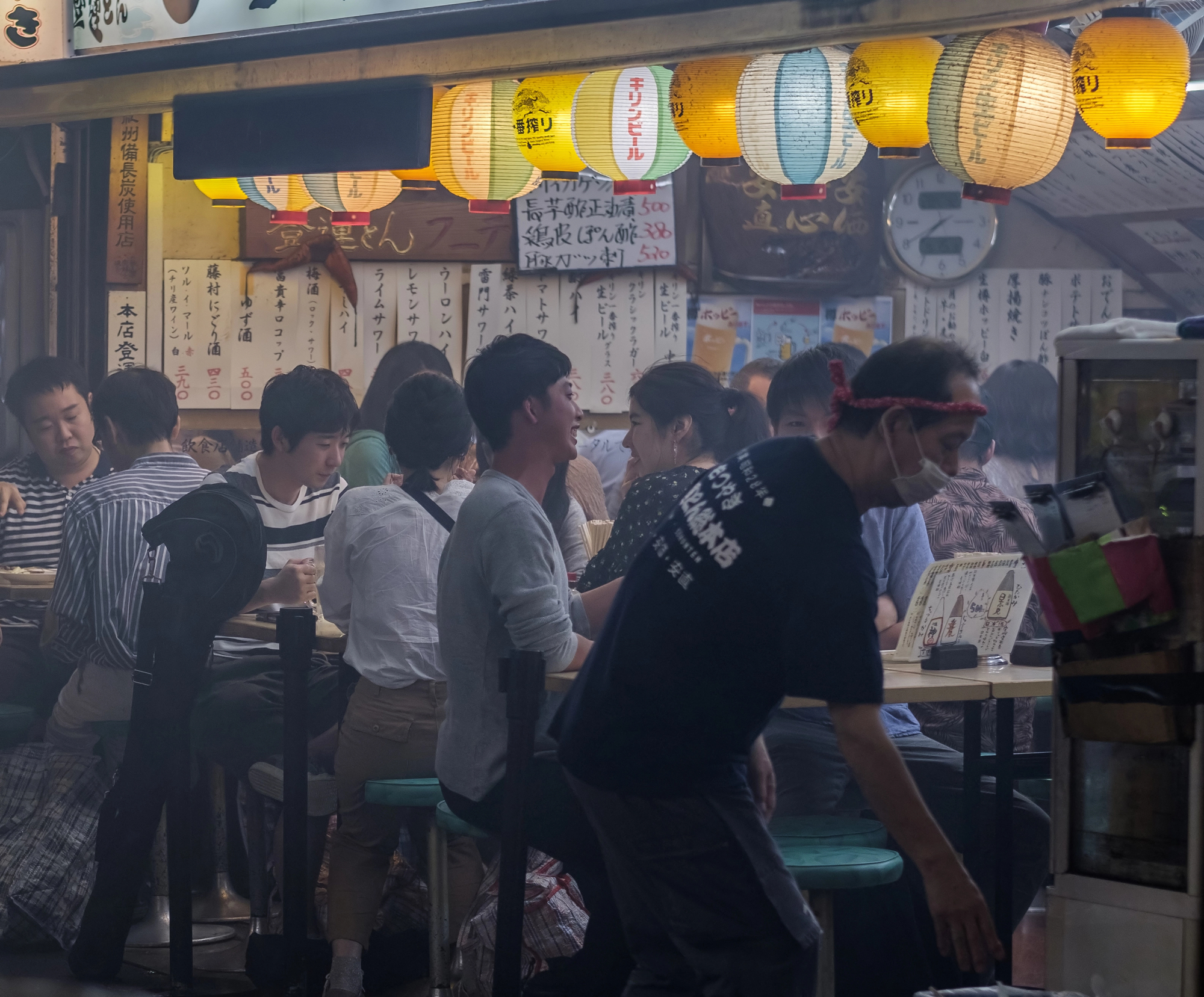 how to visit an izakaya 2 People sit and eat at crowded tables in a Japanese restaurant decorated with paper lanterns and menus written in Japanese on the walls, offering an authentic Izakaya experience reminiscent of what you might find during Japan travel.