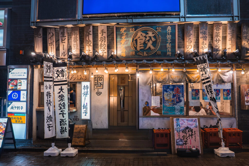 how to visit an izakaya 3 An inviting Izakaya, this Japanese restaurant features illuminated signs, hanging banners with Japanese text, and colorful posters at the entrance—all reflected on the wet street at night. If you visit Japan, this spot promises an authentic atmosphere and warm hospitality.