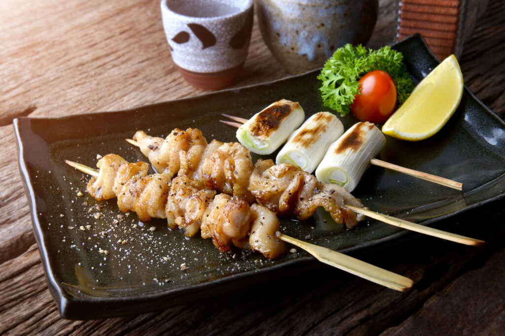 izakaya food kushiyaki Two skewers of grilled chicken, a skewer of grilled green onions, cherry tomato, parsley, and a lemon wedge are artfully arranged on a black plate next to ceramic cups on a rustic wooden surface—perfect for those seeking an authentic Izakaya experience reminiscent of Japan.