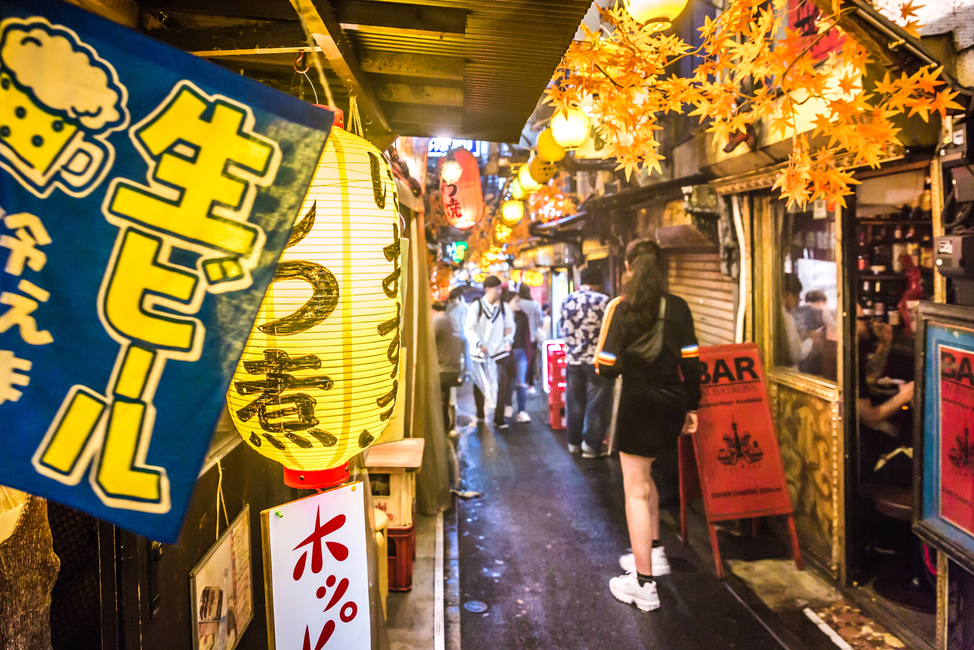 izakaya in alley in tokyo japan Narrow alley lined with Japanese lanterns, bar signs, and autumn leaves decorations; people walk and gather in the vibrant nightlife scene, discovering hidden Japanese pubs perfect for anyone looking to visit Izakaya Japan.