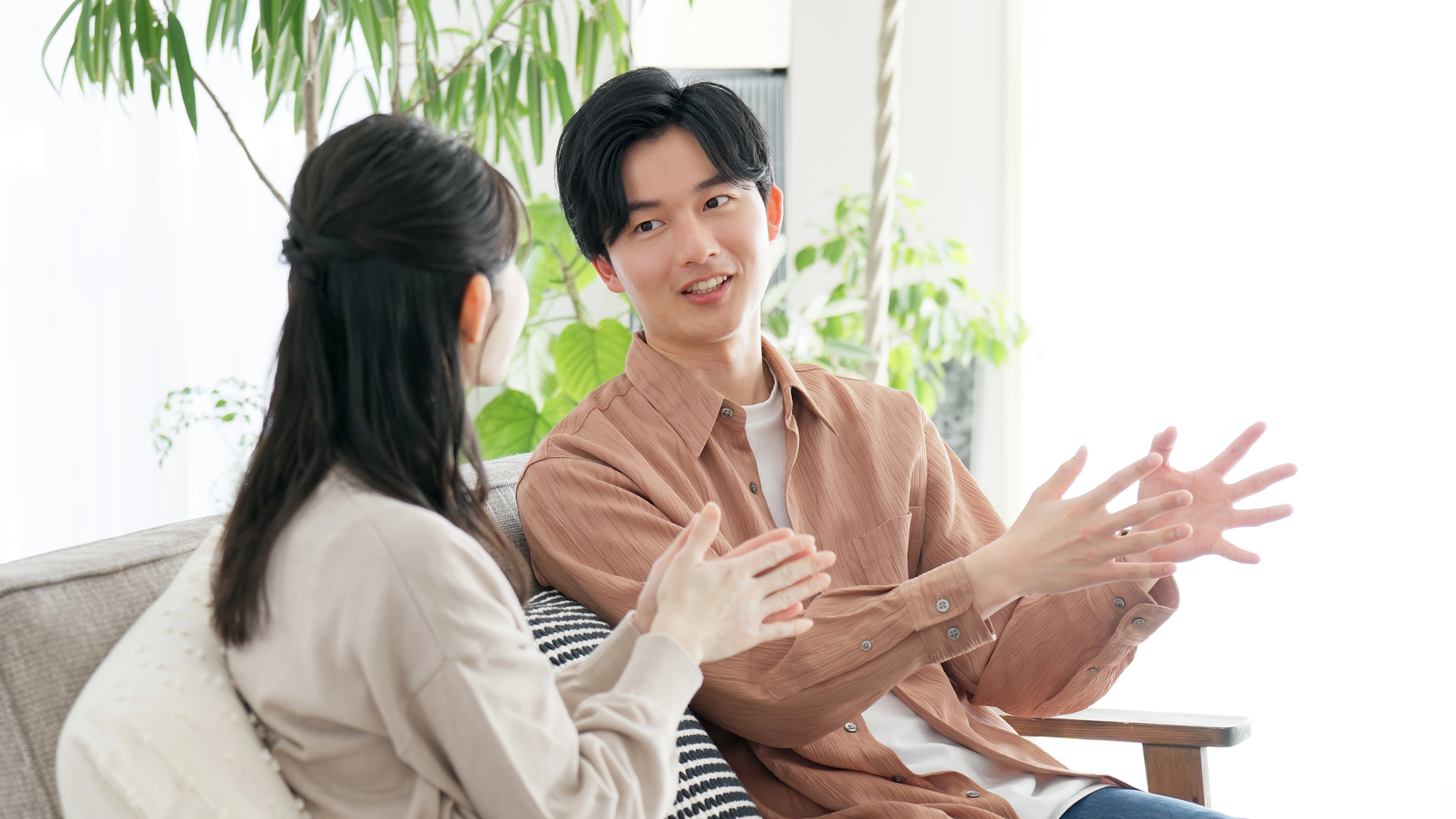 japanese filler words 2 Two people sit on a couch indoors, engaged in conversation with expressive hand gestures as they casually use Japanese filler words. Green plants are visible in the background
