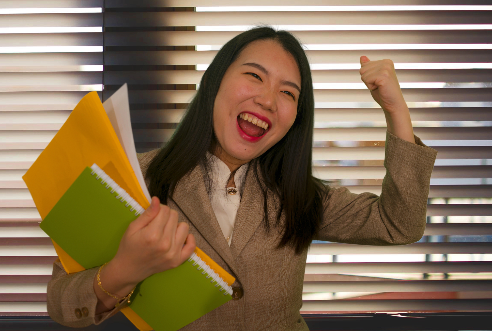 A woman in a suit holding folders and a notebook smiles and raises her fist in excitement, embodying the energy of “I can't wait!”