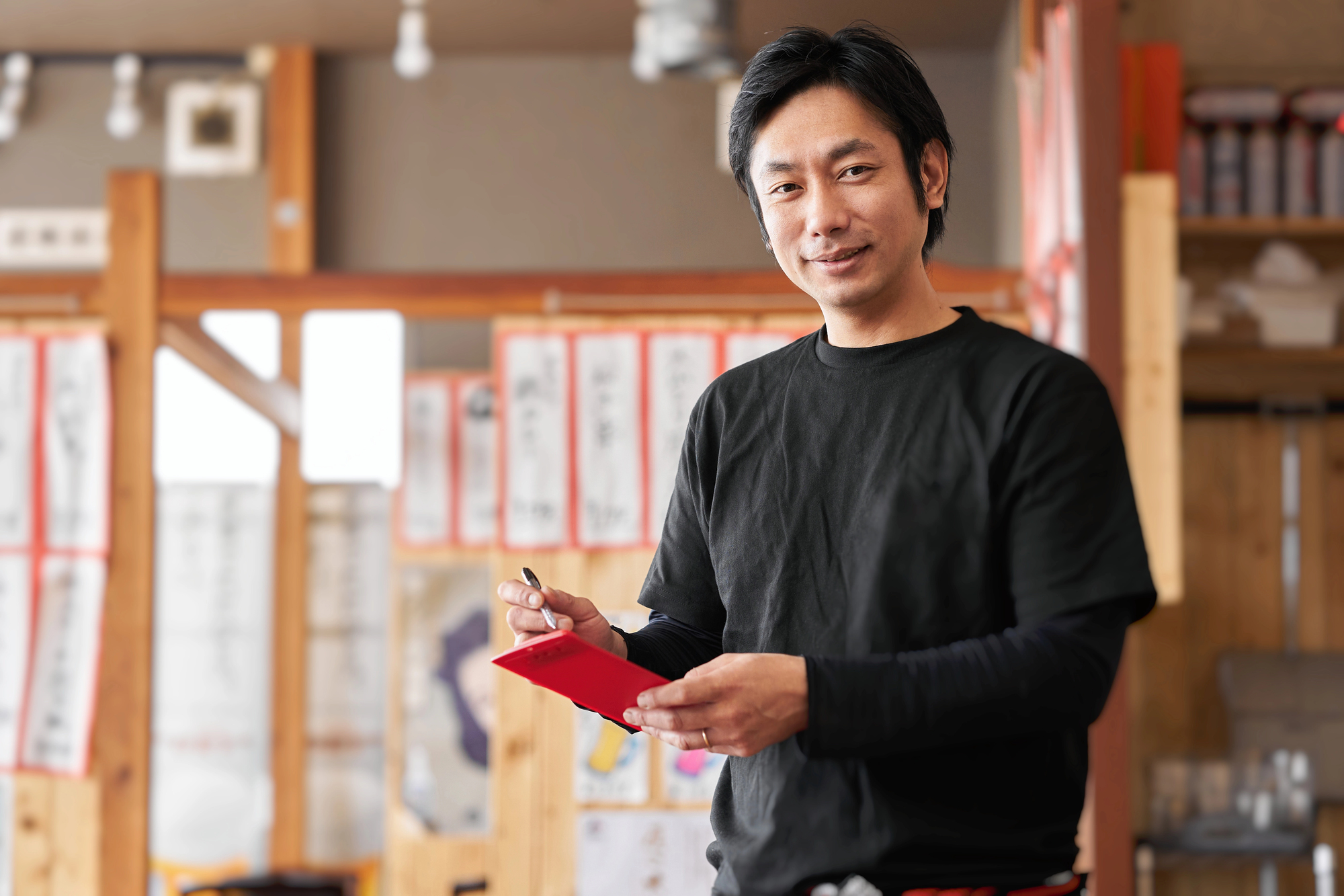 Man in a black shirt holding a notepad and pen, standing in a restaurant with wooden decor and Japanese signs on the walls, as if ready to take an order or politely ask you to “wait” in Japanese.