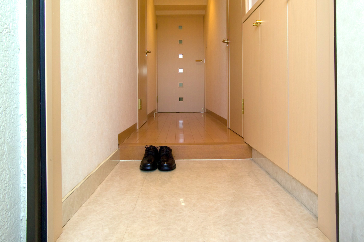 A pair of black shoes is placed neatly at the entrance of a narrow, brightly lit hallway with cream-colored walls and doors, reflecting Japanese home etiquette.
