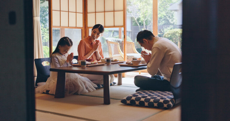 A Japanese family sits on the tatami floor around a low table in a sunlit room, hands clasped before a meal.
