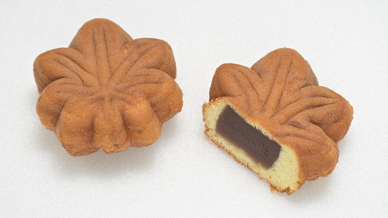 Two momiji manju - maple leaf-shaped cakes - are shown, one whole and one cut in half to reveal a dark bean paste filling inside—a treat often enjoyed during October in Japan.