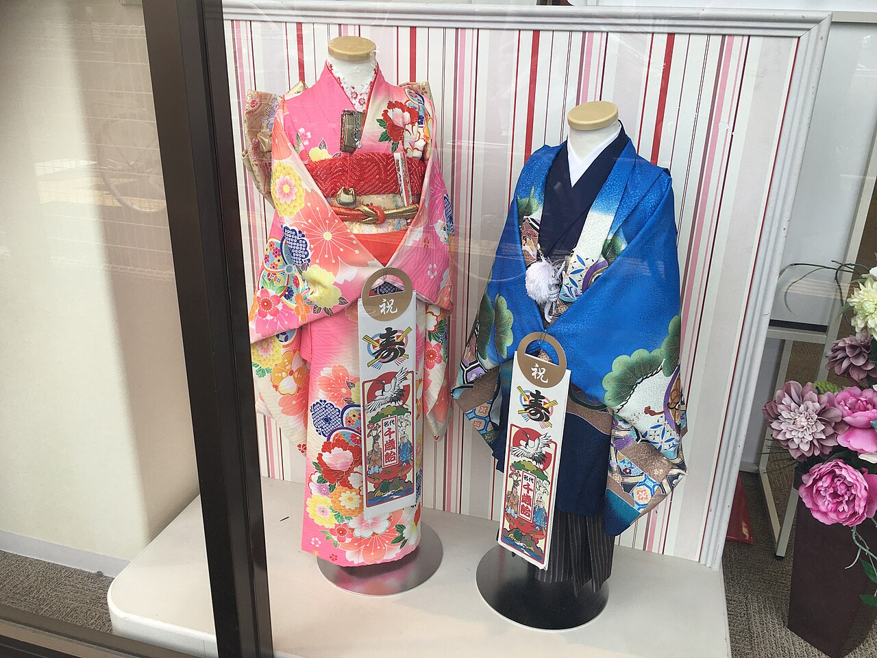 Two mannequins display traditional Japanese children’s kimonos, one pink and one blue, behind a glass window with striped and floral decorations.