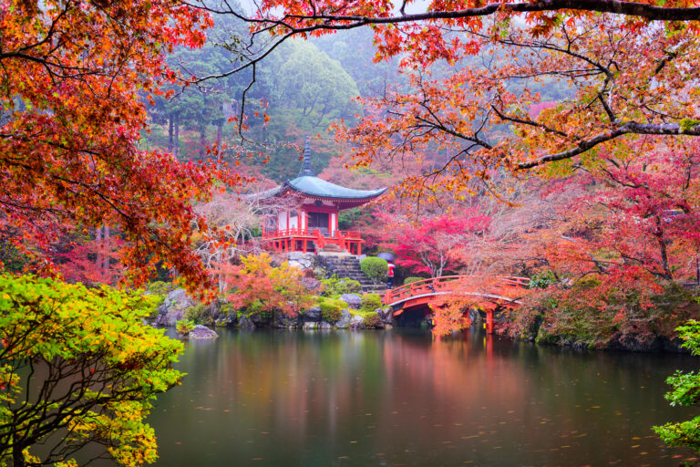 A red-painted Japanese temple framed by trees and foliage in bright autumnal colours of red, orange and gold.